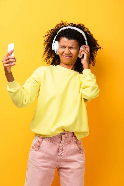 Displeased african american woman in headphones holding smartphone on yellow — Stock Photo
