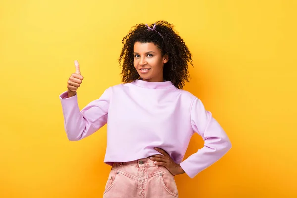 Feliz afroamericana mujer de pie con la mano en la cadera y mostrando el pulgar hacia arriba en amarillo — Stock Photo