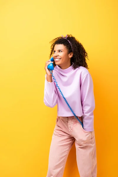 Happy african american woman talking on blue retro phone on yellow — Stock Photo