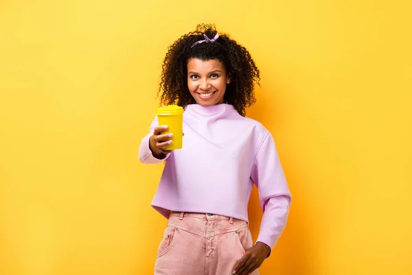 Gaie femme afro-américaine tenant thermotasse et debout avec la main dans la poche sur jaune — Photo de stock