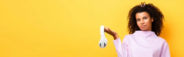 Mulher africana americana segurando fones de ouvido sem fio em amarelo, banner — Stock Photo