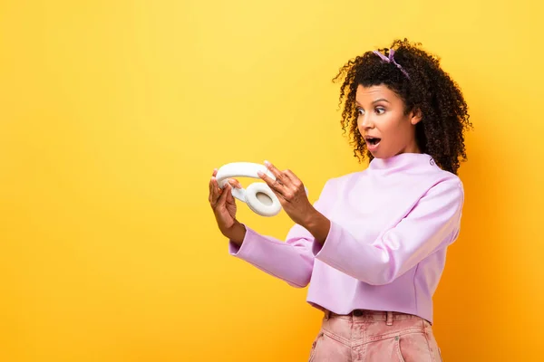 Femme afro-américaine choquée tenant écouteurs sans fil sur jaune — Photo de stock