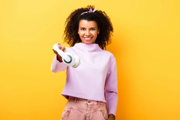 Feliz mulher afro-americana segurando fones de ouvido sem fio no amarelo — Fotografia de Stock