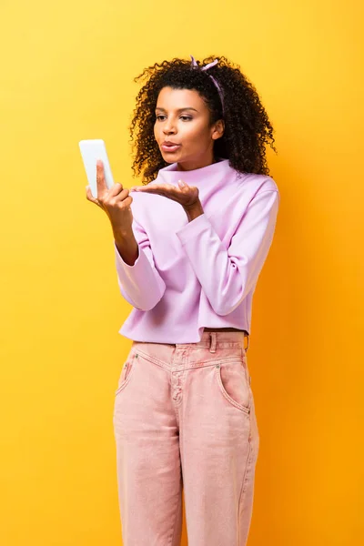 African american woman holding smartphone and sending air kiss on yellow — Stock Photo