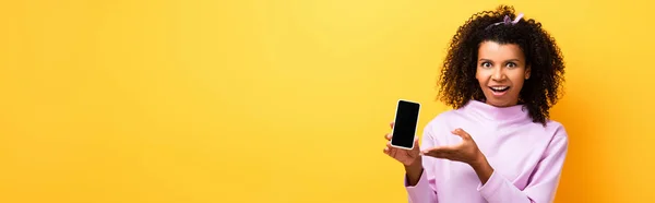 Donna afroamericana eccitata che punta con mano lo smartphone con schermo bianco su giallo, banner — Foto stock