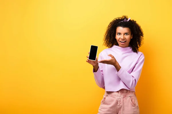 Aufgeregte Afroamerikanerin zeigt mit der Hand auf Smartphone mit leerem Bildschirm auf gelb — Stockfoto