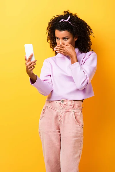 Chocado Africano americano mulher cobrindo boca enquanto olha para smartphone no amarelo — Fotografia de Stock