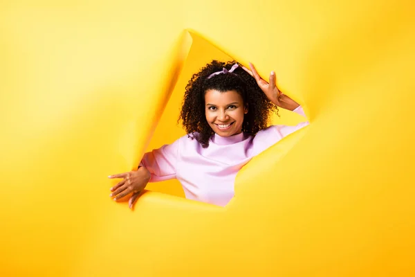 Happy african american woman looking at camera through hole in ripped paper on yellow background — Stock Photo