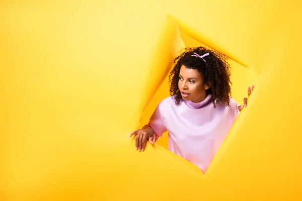 Curious african american woman looking away through hole in ripped paper on yellow background — Stock Photo