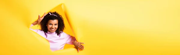 Happy african american woman looking at camera through hole in ripped paper on yellow background, banner — Stock Photo