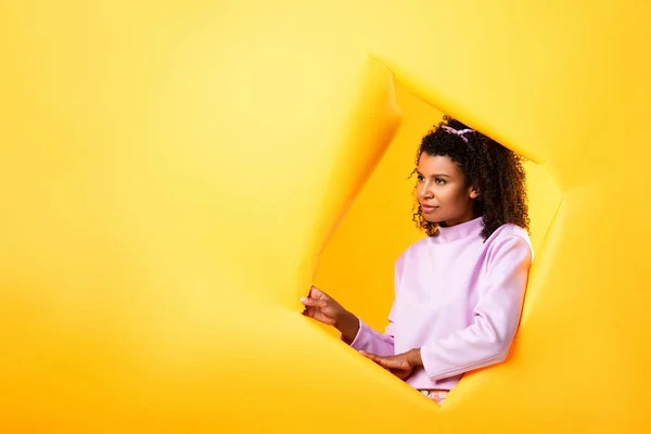 African american woman looking away through hole in ripped paper on yellow background — Stock Photo