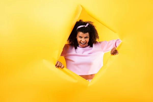 Arrabbiato africano americano donna guardando fotocamera attraverso buco in carta strappata su sfondo giallo — Foto stock