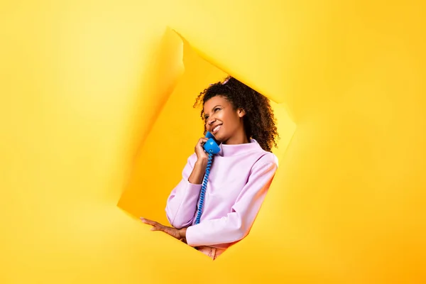 Feliz mulher afro-americana falando no telefone retro perto buraco em papel rasgado no fundo amarelo — Fotografia de Stock