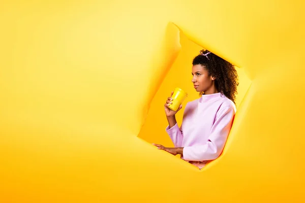 Mujer afroamericana sosteniendo eco taza cerca de agujero en papel rasgado sobre fondo amarillo - foto de stock