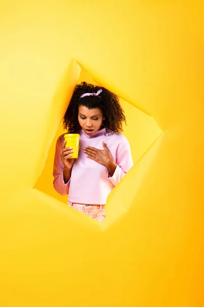 Überrascht afrikanisch-amerikanische Frau mit Einwegbecher und Blick nach unten durch ein Loch in zerrissenem Papier auf gelbem Hintergrund — Stockfoto