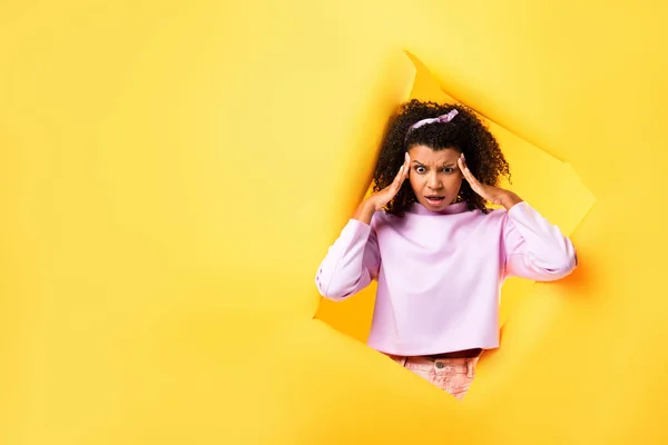 Shocked african american woman touching temples and looking down near hole in ripped paper on yellow background — Stock Photo