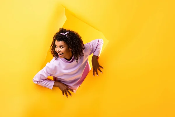 Curious african american woman looking away through hole in ripped paper and smiling on yellow background — Stock Photo