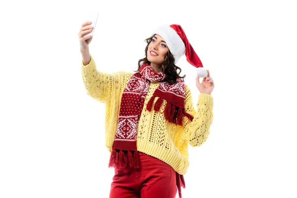 Радостная женщина в шляпе Санты и шарфе, делающая селфи на смартфоне, изолированном на белом — стоковое фото