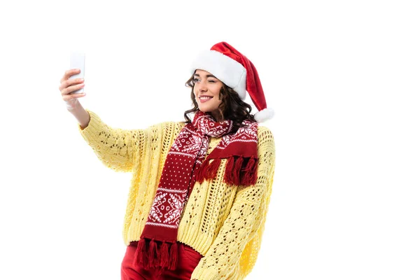 Alegre joven en sombrero de santa y bufanda tomando selfie en teléfono inteligente aislado en blanco - foto de stock