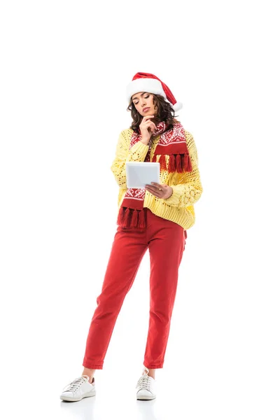 Mujer joven pensativa en sombrero de santa y bufanda mirando tableta digital aislada en blanco - foto de stock