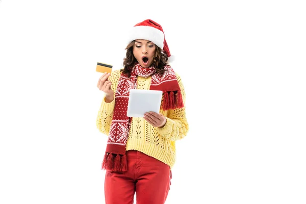 Schockierte junge Frau mit Weihnachtsmütze und Schal schaut auf digitales Tablet, während sie ihre Kreditkarte isoliert auf weiß hält — Stockfoto