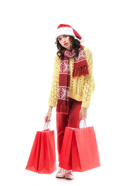 Fatigué jeune femme dans santa chapeau et écharpe avec ornement tenant sacs à provisions rouges isolés sur blanc — Photo de stock