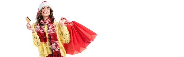 Concepto panorámico de mujer complacida en sombrero de santa y bufanda con adorno sosteniendo bolsas de compras y tarjeta de crédito aislada en blanco - foto de stock