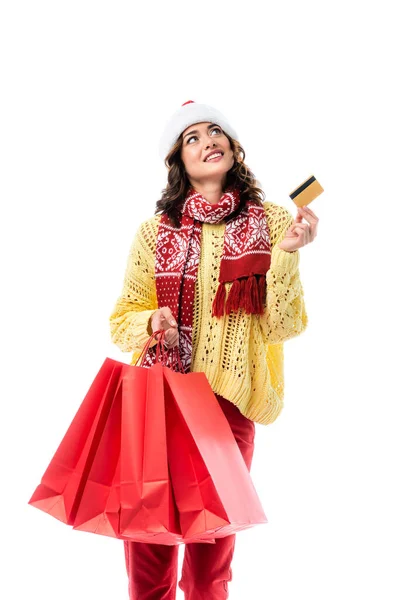 Jeune femme heureuse en santa chapeau et écharpe avec ornement tenant des sacs à provisions et carte de crédit isolé sur blanc — Photo de stock