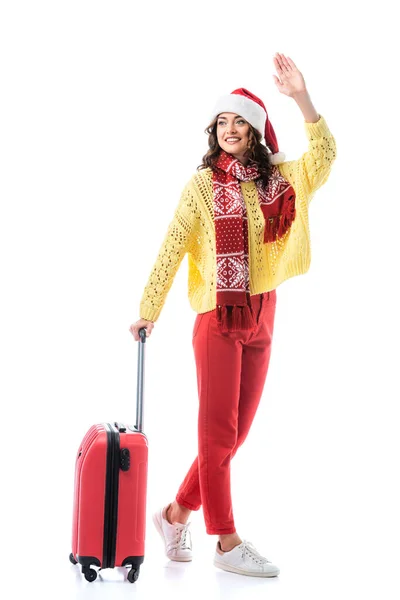 Молода жінка в капелюсі Санта та шарфі з орнаментом, що стоїть з дорожньою сумкою та махає рукою ізольовано на білому — стокове фото