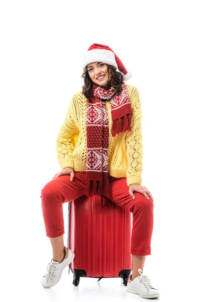 Gioiosa giovane donna in cappello di Babbo Natale e sciarpa con ornamento seduta sulla borsa da viaggio isolata su bianco — Foto stock