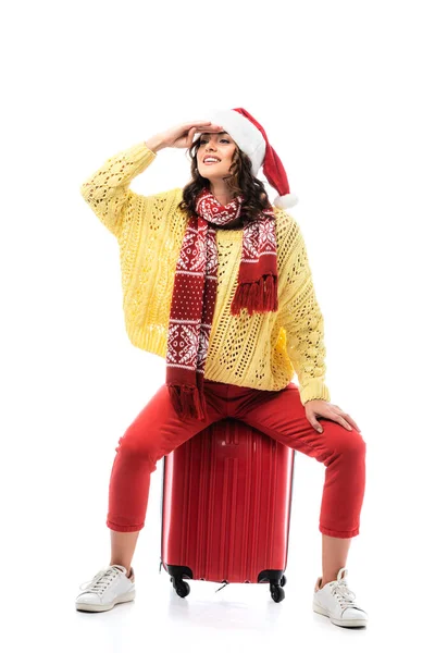 Joyeuse jeune femme en santa chapeau et écharpe avec ornement assis sur sac de voyage et regardant loin isolé sur blanc — Photo de stock