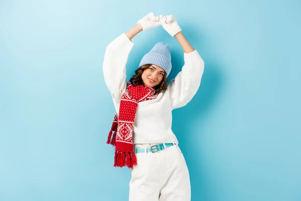 Young woman in winter outfit and accessories standing with hands above head on blue — Stock Photo