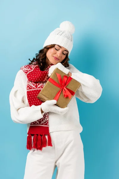 Freudige Frau mit geschlossenen Augen, im winterlichen Outfit mit umwickeltem Geschenk auf blauem Grund — Stockfoto