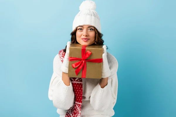 Freudige Frau im winterlichen Outfit mit eingewickeltem Geschenk und Blick in die Kamera auf blau — Stockfoto