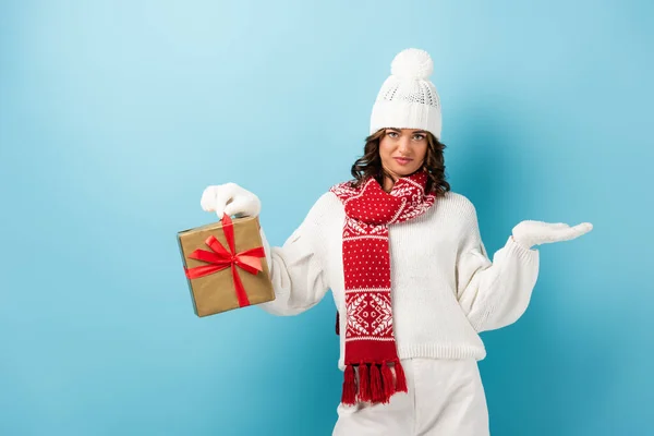 Skeptical woman in winter outfit holding wrapped present and gesturing on blue — Stock Photo