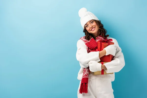 Молода задоволена жінка в білому зимовому вбранні тримає подарунки на синьому — стокове фото
