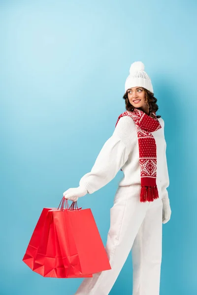 Young joyful woman in winter outfit holding red shopping bags on blue — Stock Photo