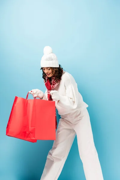 Junge fröhliche Frau im Winteroutfit blickt auf rote Einkaufstüten auf blauem Grund — Stockfoto