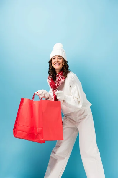 Радостная женщина в зимнем наряде, держащая красные пакеты для покупок на голубом — стоковое фото