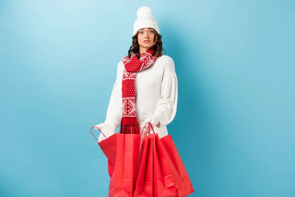 Surprised woman in winter outfit holding red shopping bags on blue — Stock Photo