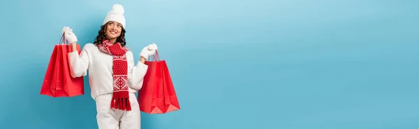 Récolte horizontale de femme joyeuse en tenue d'hiver tenant des sacs à provisions rouges sur bleu — Photo de stock