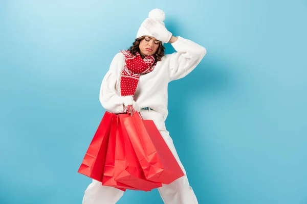 Disappointed young woman in winter outfit holding red shopping bags on blue — Stock Photo