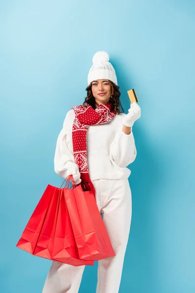 Jeune femme en tenue d'hiver tenant des sacs à provisions rouges et carte de crédit tout en clignant des yeux sur le bleu — Photo de stock