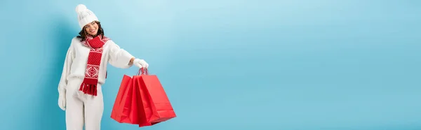 Horizontal crop of happy woman in winter outfit holding red shopping bags and looking away on blue — Stock Photo