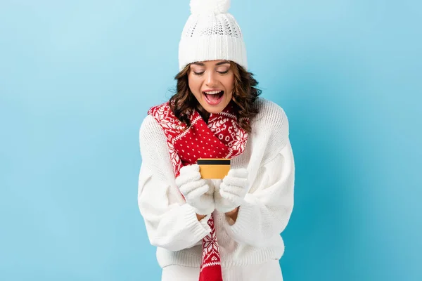 Excited young woman in winter outfit holding credit card on blue — Stock Photo