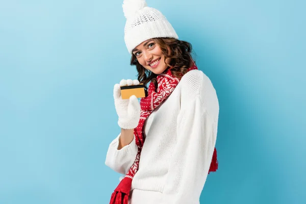 Happy young woman in white winter outfit holding credit card on blue — Stock Photo