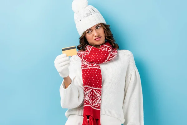Незадоволена молода жінка в білому зимовому вбранні тримає кредитну картку на синьому — стокове фото