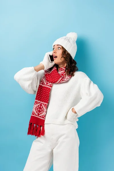 Schockierte junge Frau im weißen Winteroutfit spricht auf Smartphone auf blau — Stockfoto