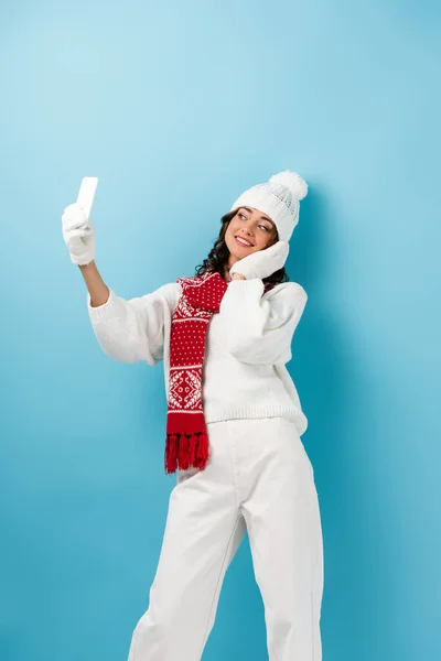 Joven mujer complacida en traje de invierno blanco tomando selfie en azul - foto de stock