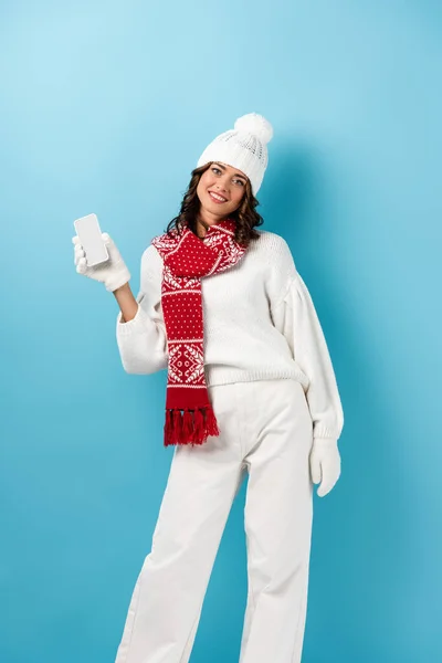 Щаслива молода жінка в зимовому вбранні тримає мобільний телефон на синьому — Stock Photo